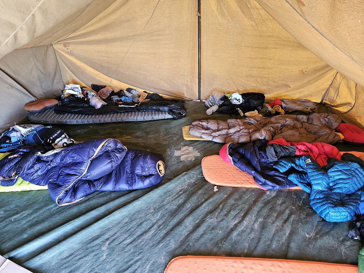Inside a multi-stage race tent, sleeping bags spread across the floor, kit piled at the edges
