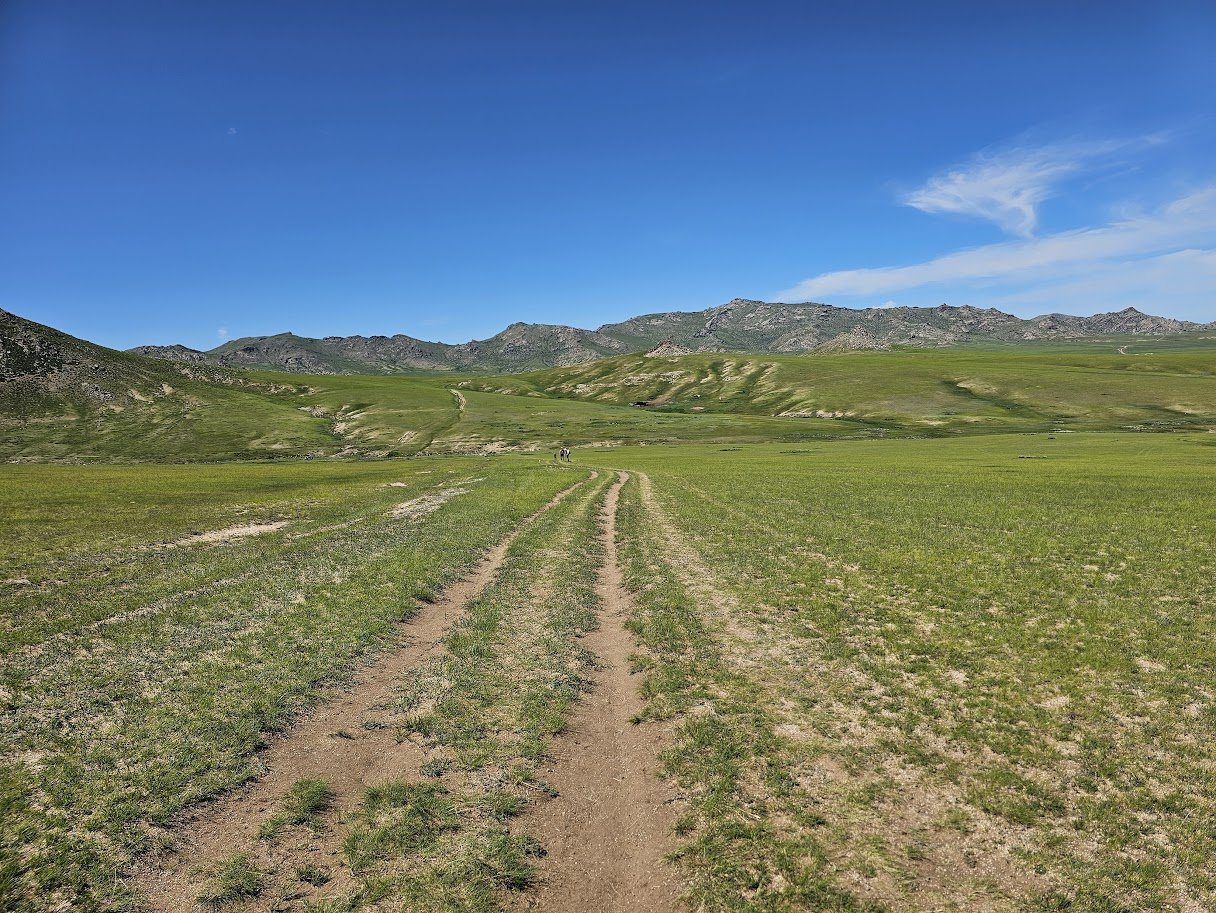 Endless rolling grassland of the Mongolian steppe with a dirt track winding into the distance and small figures of runners ahead